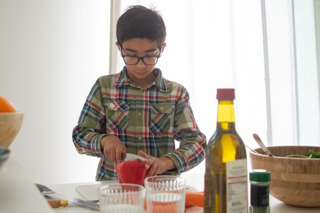 A young boy cutting red bell pepper in a bright, indoor kitchen.